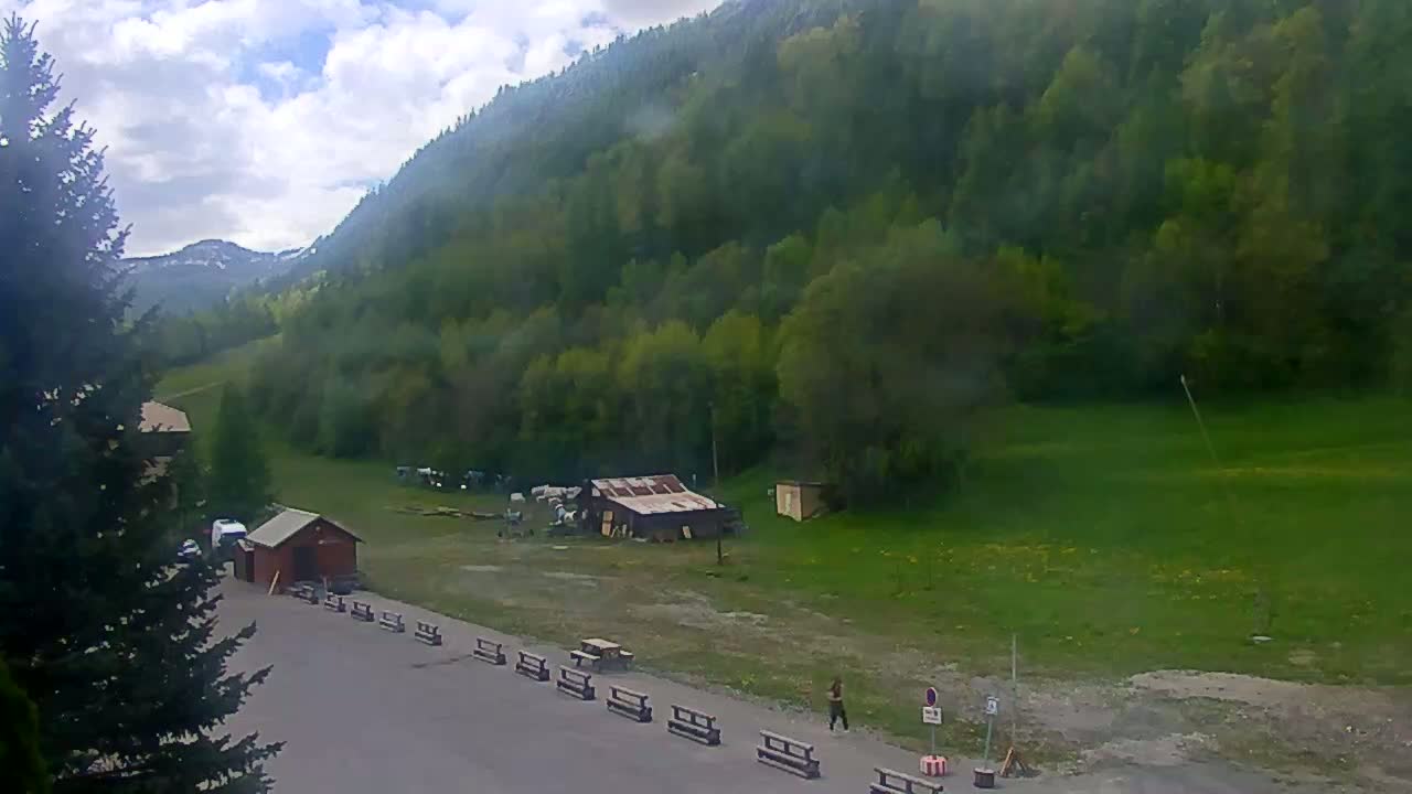 A partly cloudy day reveals a mountainous landscape with lush green forests, a few small buildings, and a person walking near a row of benches in a paved area.