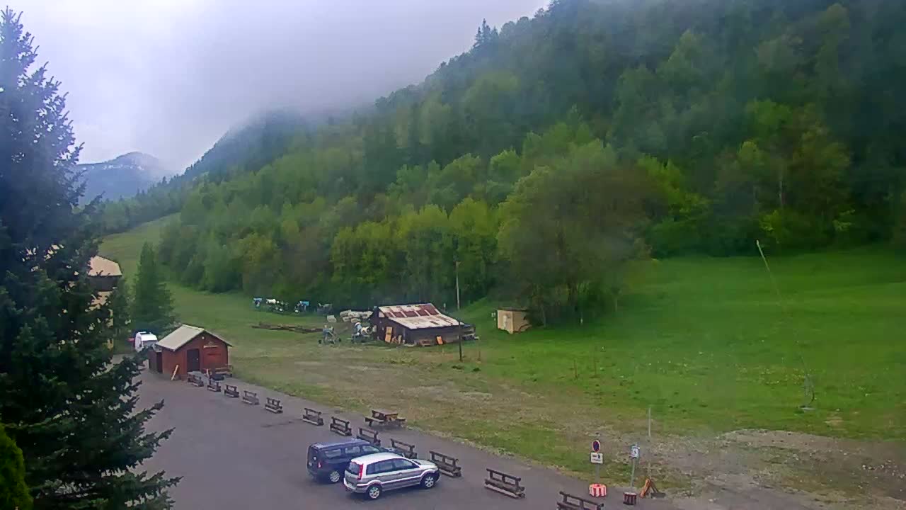 A parking lot with two cars sits before a small building and a grassy hillside covered in lush green trees under a cloudy sky.