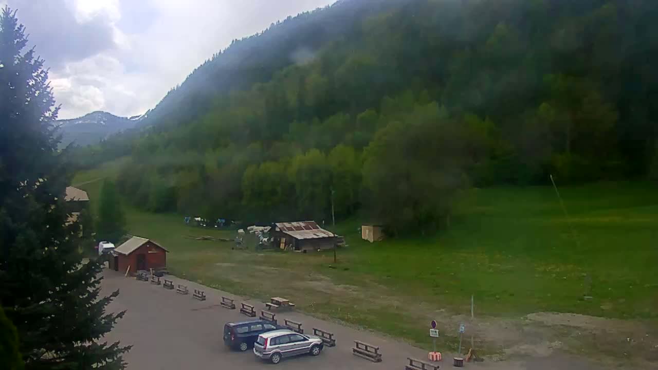 A cloudy day reveals a mountainside covered in lush green trees, with a small parking lot containing a couple of cars and some simple buildings below.