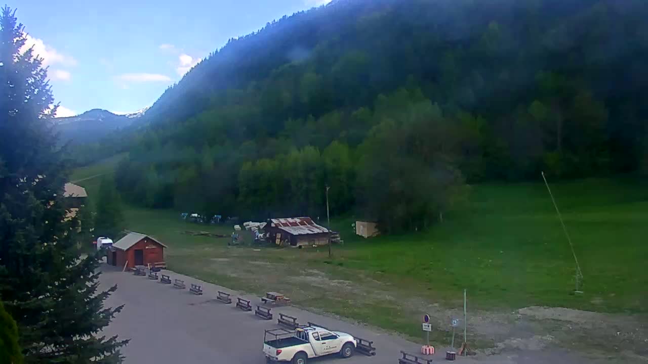 A mostly clear day shows a mountainous landscape with green trees and grass, a small parking area with a few buildings and a white pickup truck.