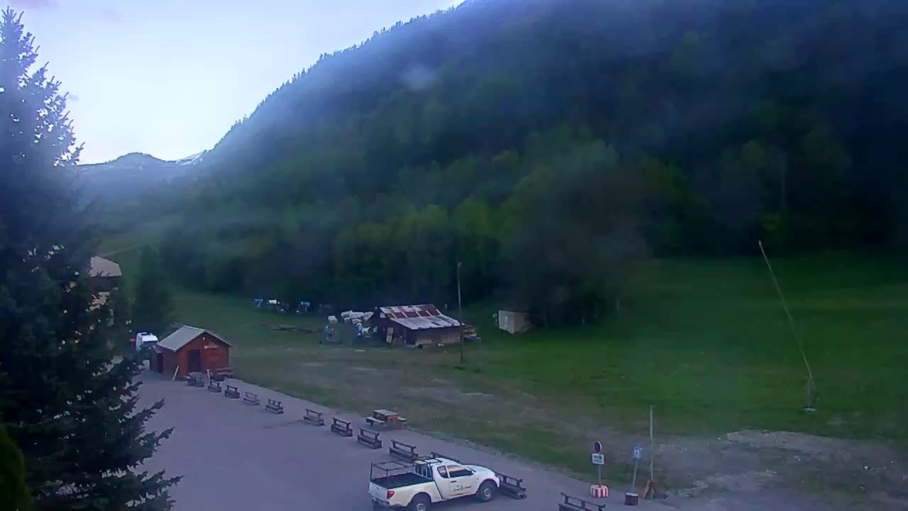 A high-angle, long shot shows a green, grassy hillside under a hazy sky, with a small parking lot containing a pickup truck and some small buildings at the base of the hill.