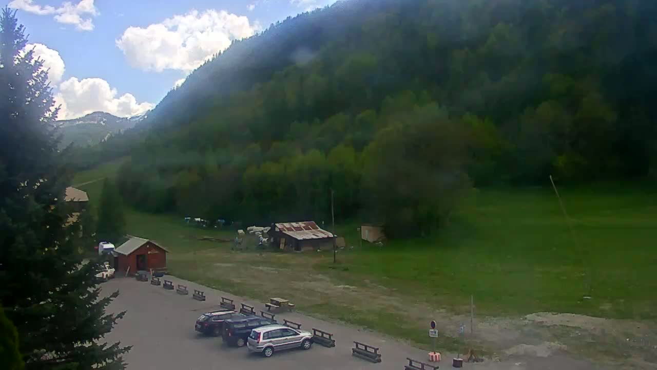 A partly cloudy day reveals a mountainside covered in lush green trees, a small parking lot with several cars, and a few simple buildings near the base of the mountain.