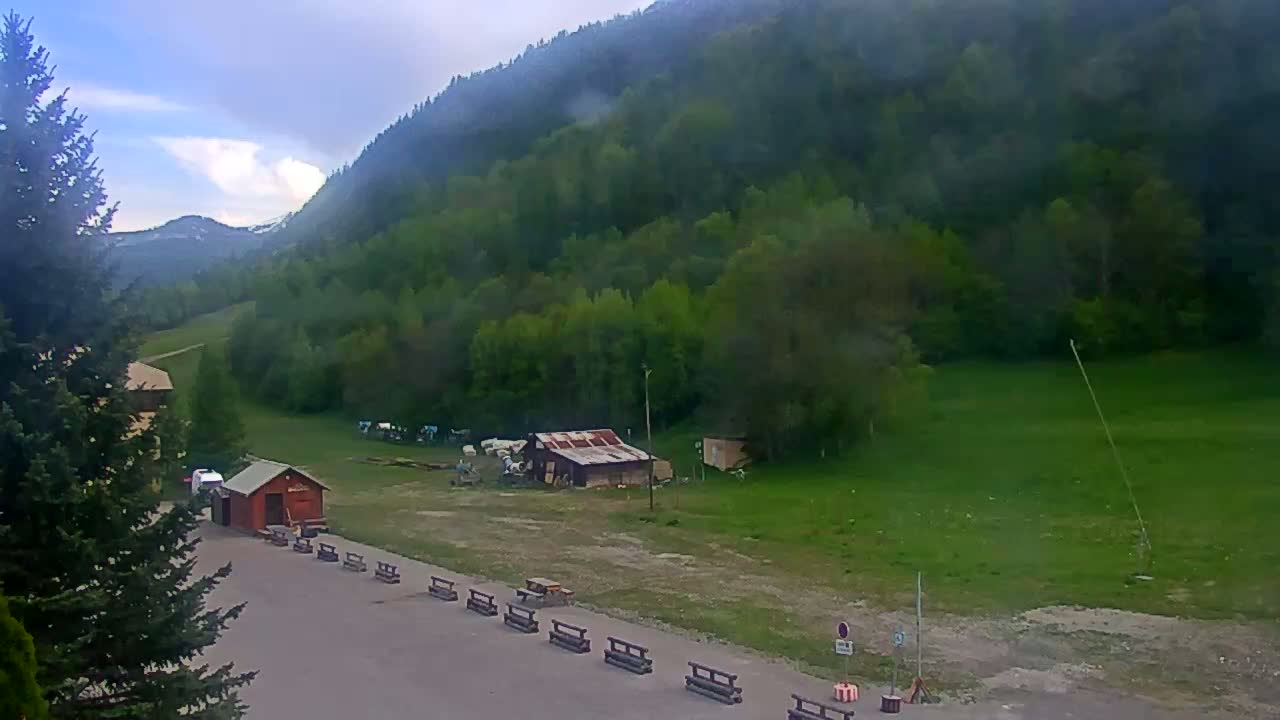 A mostly cloudy day reveals a mountainous landscape with green trees and fields, a small red building, and a paved area with several benches.