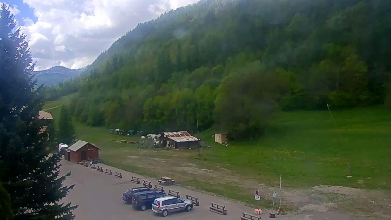 A parking lot with a few cars sits below a mountain covered in lush green trees under a partly cloudy sky.