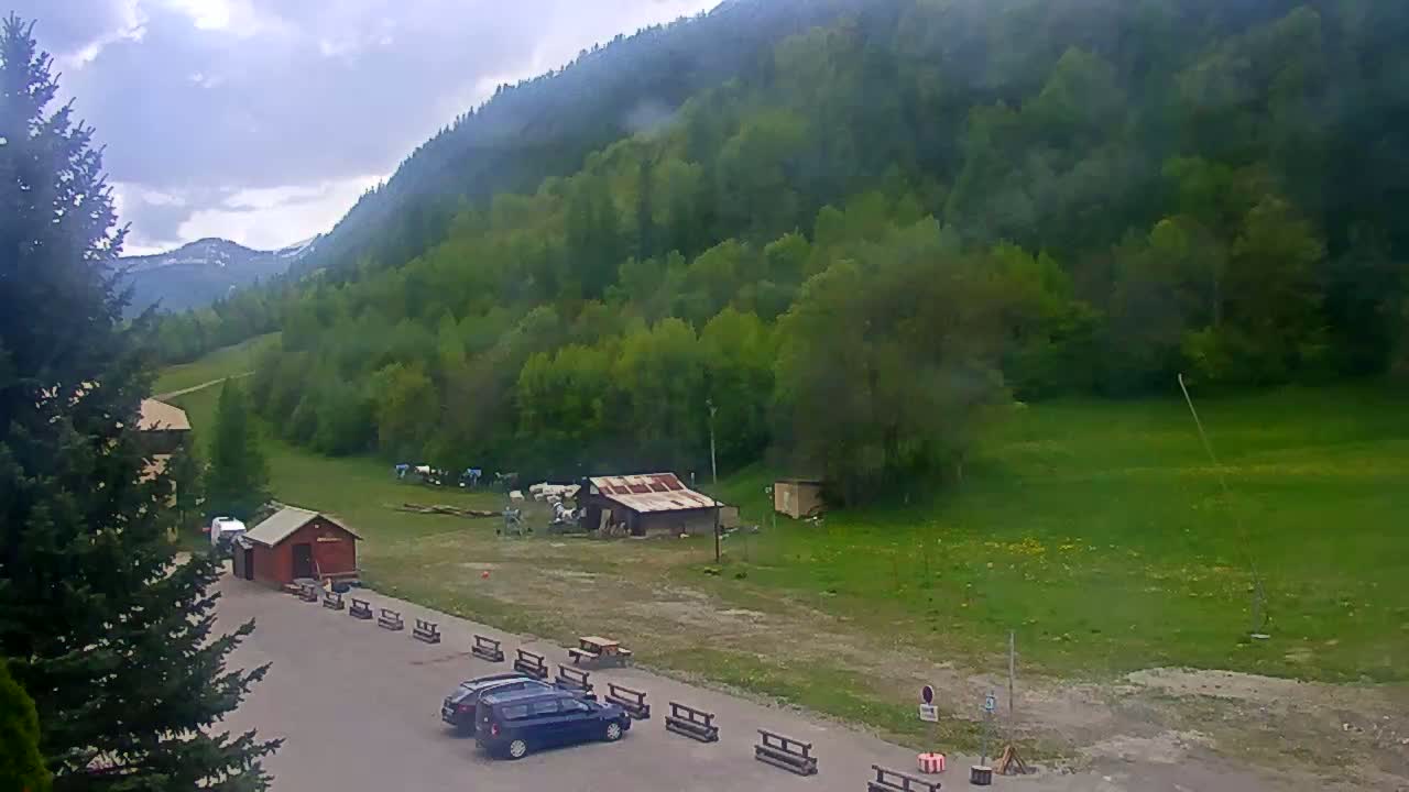 A parking lot with a few cars sits before a grassy field and a lush green mountainside under a cloudy sky.