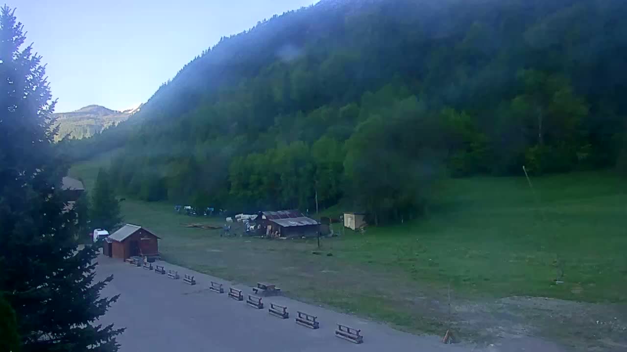 A hazy, clear morning shows a mountainside covered in lush green trees, a flat grassy area with a few small buildings and picnic tables, and a paved area in the foreground near a tall evergreen tree.