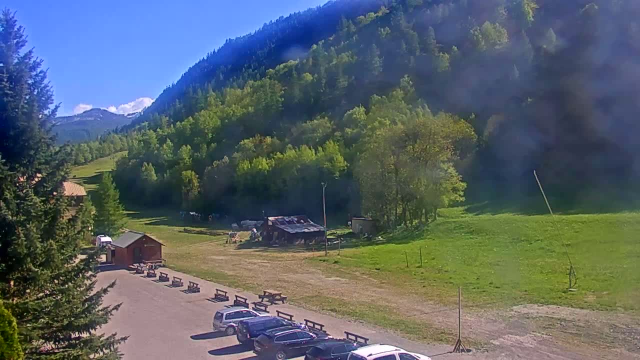 A parking lot with several cars is situated in front of a small building, overlooking a grassy field and a verdant mountainside under a clear, sunny sky.