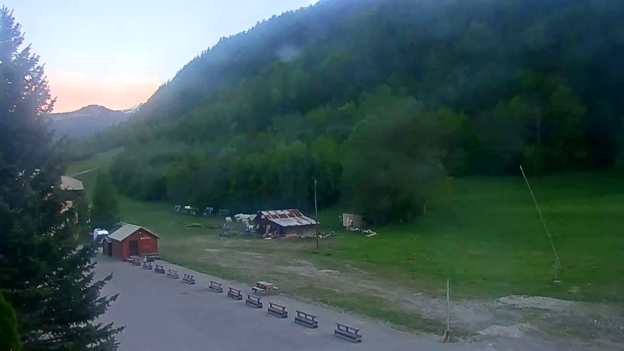 A small, red building sits in a grassy area near a mountain slope under a hazy, pinkish sunrise.