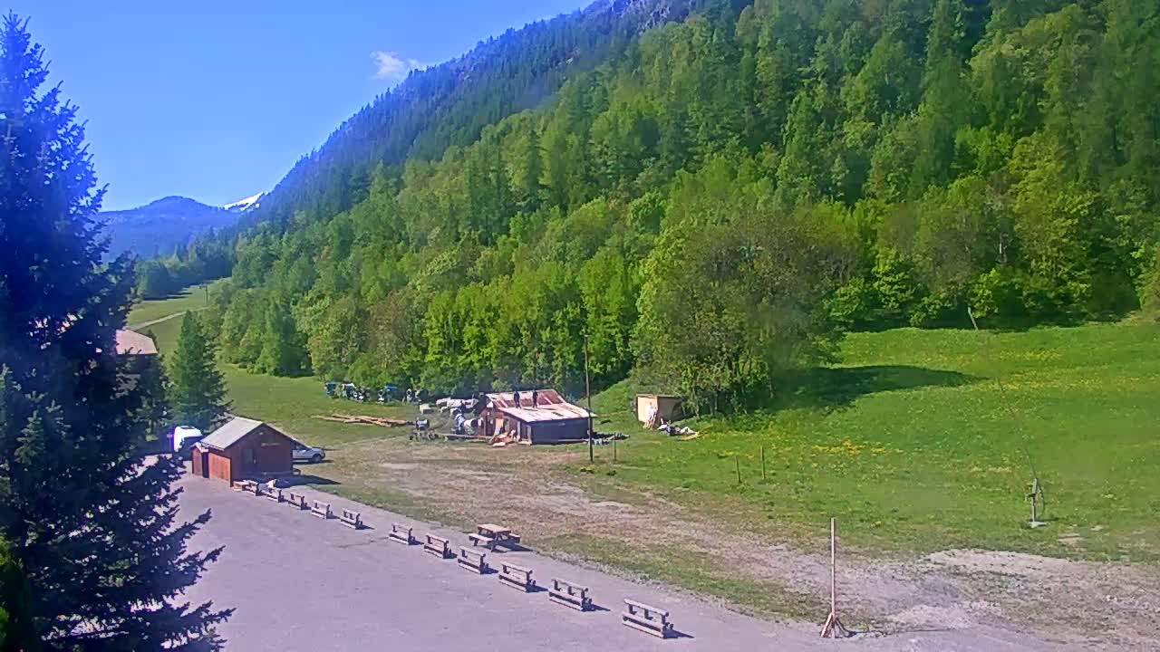 A sunny mountain valley scene features a small building under construction, several parked vehicles, picnic tables, and a grassy field next to a lush green forest.