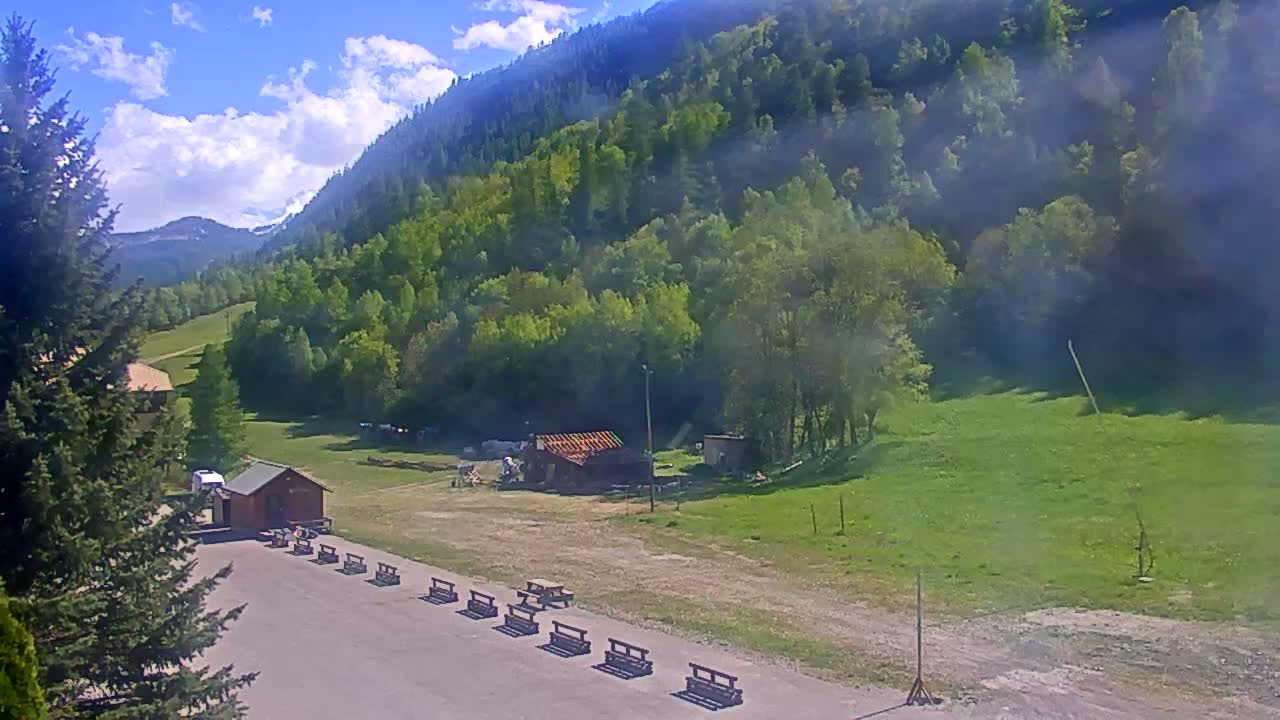 A sunny day in a mountainous region shows a grassy field with a small building and a row of benches, next to a parking lot and surrounded by lush green forests.