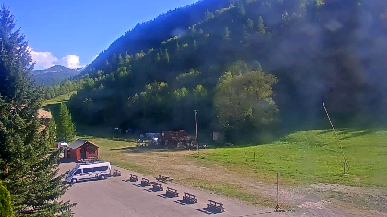 A white van is parked in a paved area next to several empty benches, overlooking a grassy field and a verdant mountainside under a clear blue sky.