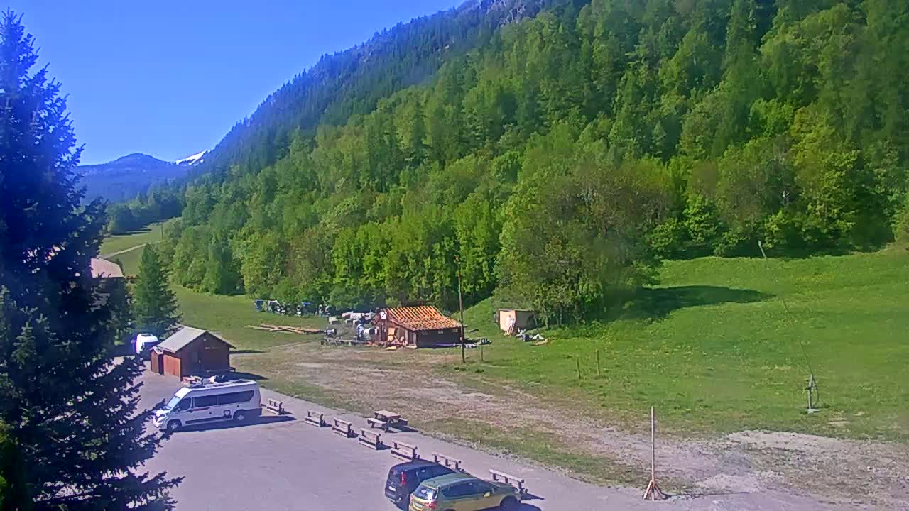 A sunny day reveals a mountainside covered in lush green trees, a small building, and a parking lot with several vehicles, all situated near a grassy field.
