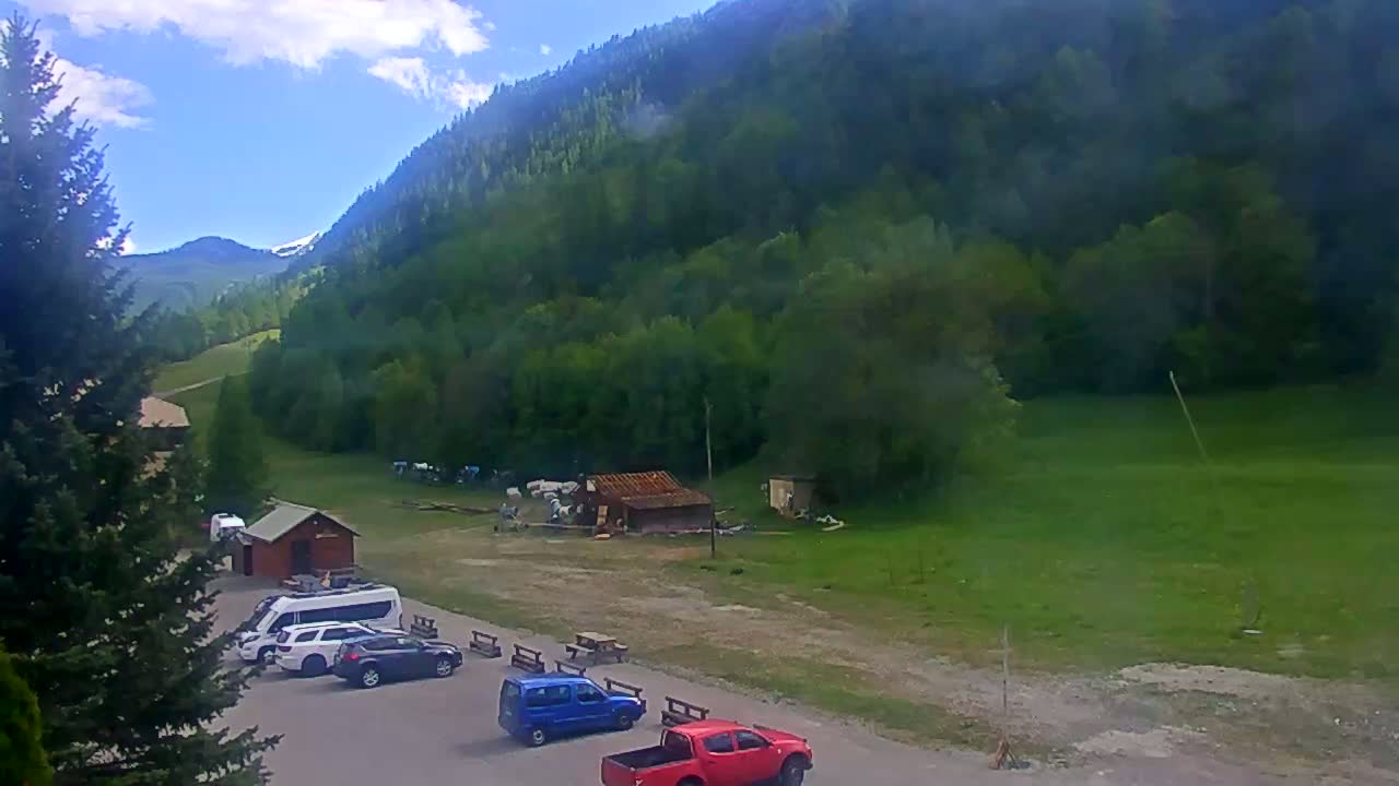 A mountainside parking lot with several cars and vans is nestled below a lush green mountain under a partly cloudy sky.