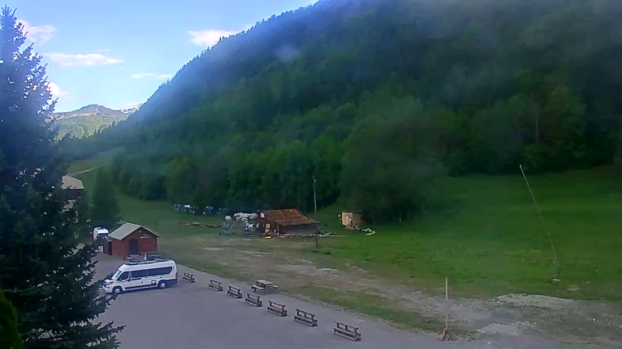 A white van is parked in a mountain valley under a mostly clear sky, near a small building and several picnic tables.