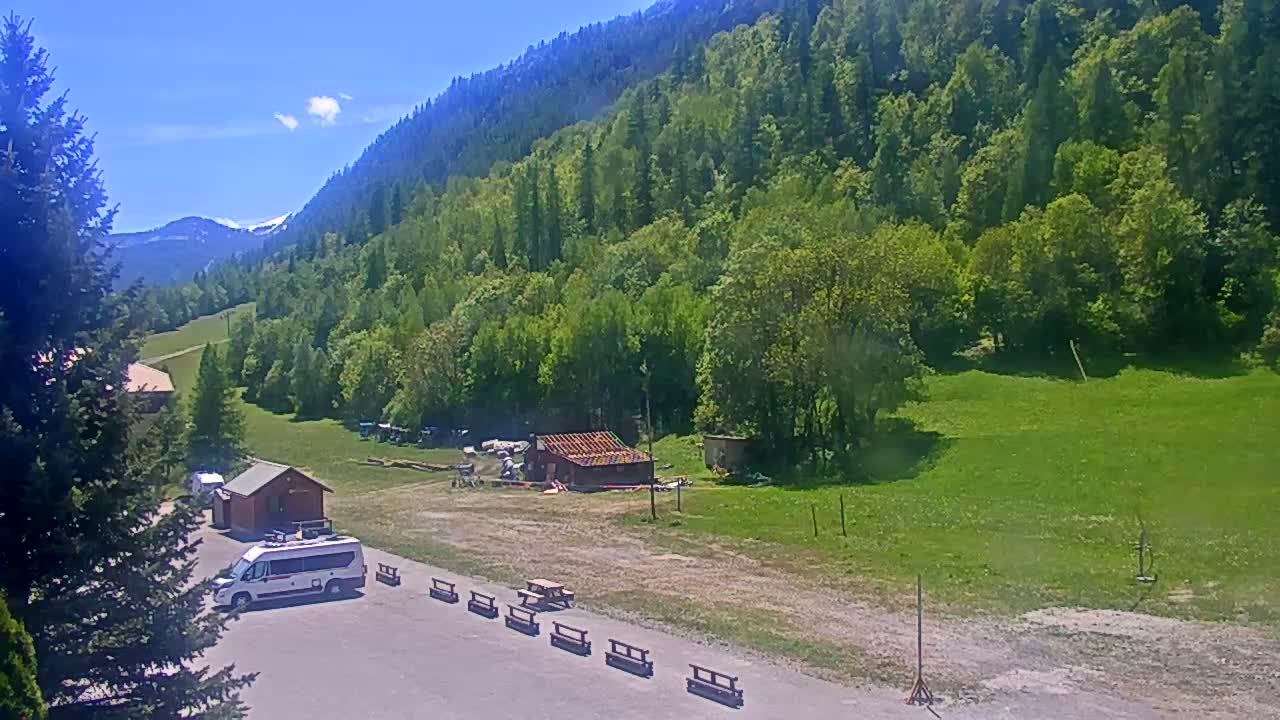 A van is parked in a clearing next to a small building, surrounded by lush green fields and a mountainside covered in dense forest under a mostly sunny sky.