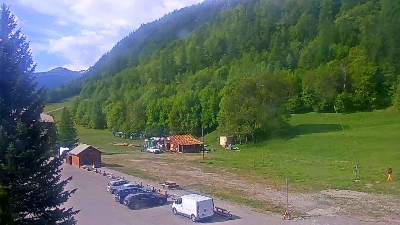 A parking lot with several cars and a van is situated below a lush green mountainside on a partly cloudy day.