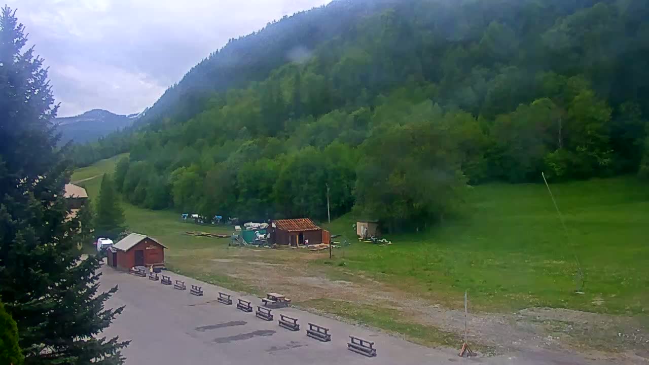 A view of a grassy area with a few small buildings and picnic tables, situated below a lush green mountainside on an overcast day.