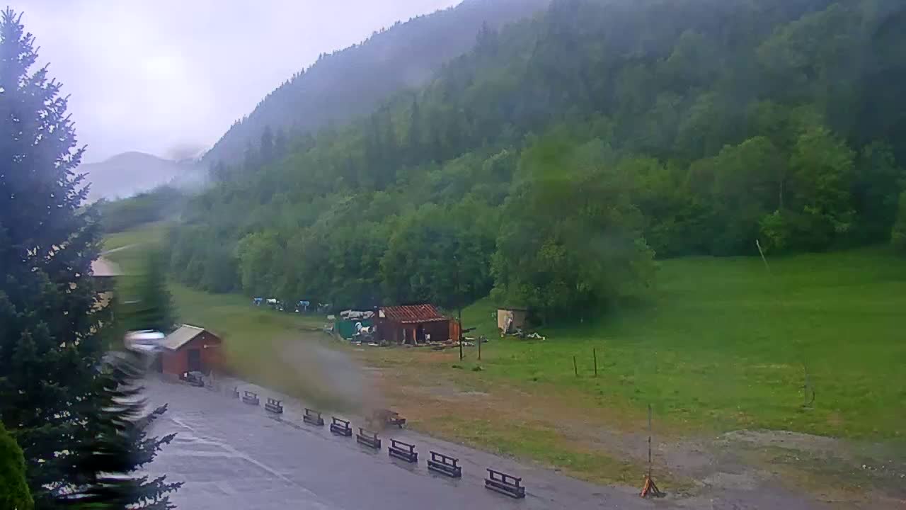 A rainy view of a green hillside with a few small buildings and a roadway in the foreground.