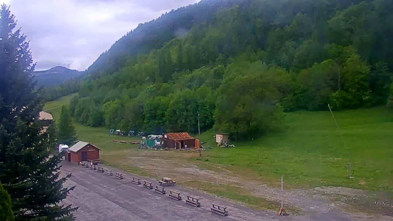 A mostly cloudy day shows a grassy area with a few small buildings and picnic tables nestled at the base of a lush green mountain.
