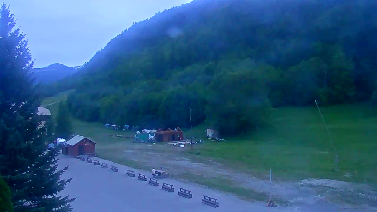A mountainside clearing under an overcast sky shows a small building under construction, a few parked vehicles, and several wooden benches lined up along a paved area.