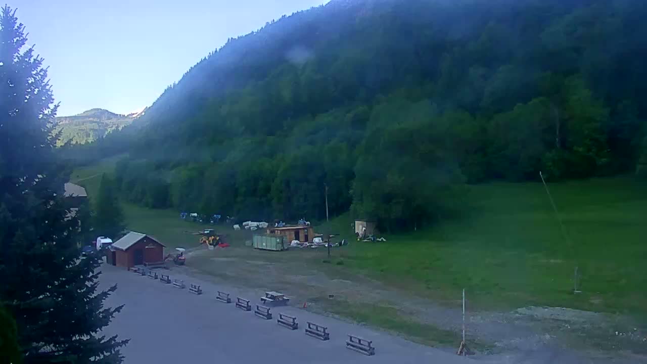 A hazy, clear morning reveals a mountainside covered in lush green trees, a flat area below with small buildings, construction equipment, and picnic tables, and a paved road in the foreground.
