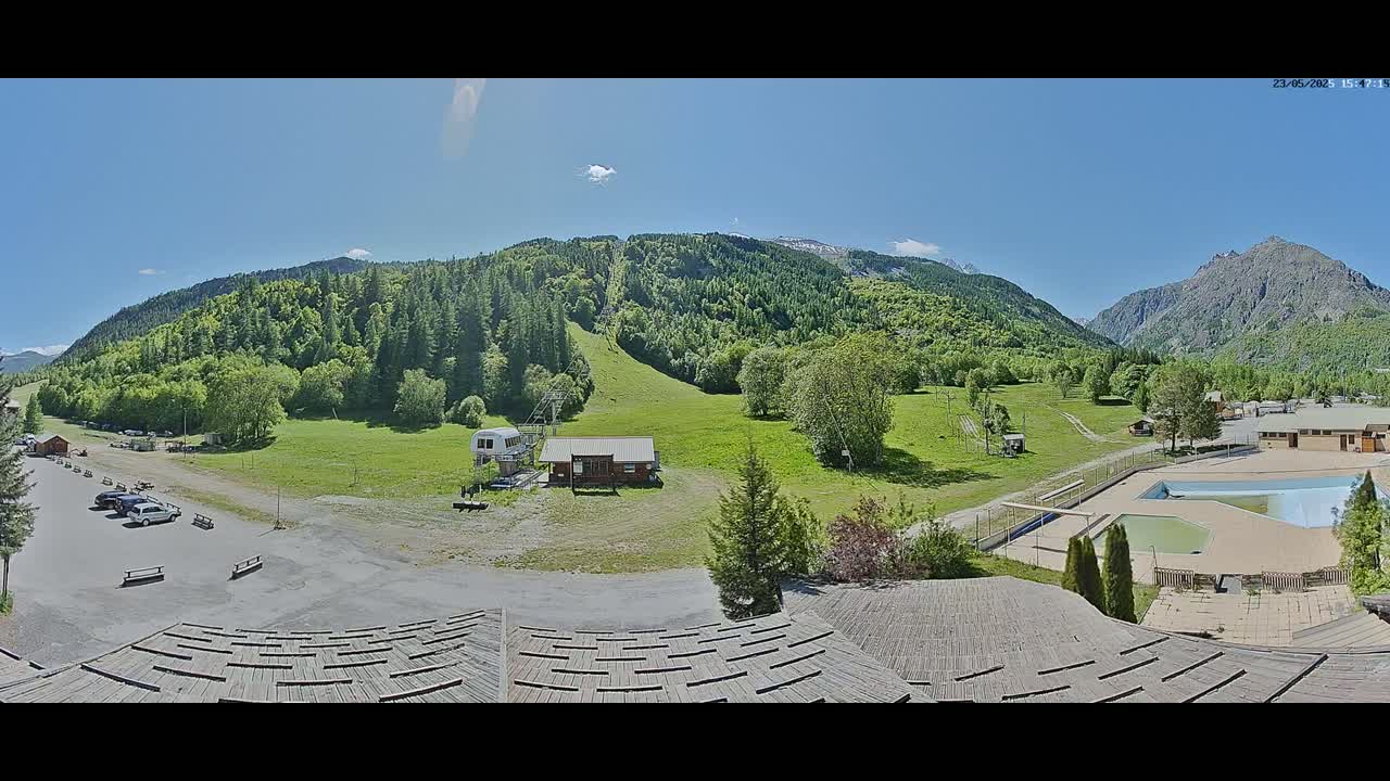 A sunny mountain valley scene with a ski lift, small buildings, a parking lot, and a swimming pool, viewed from a rooftop.