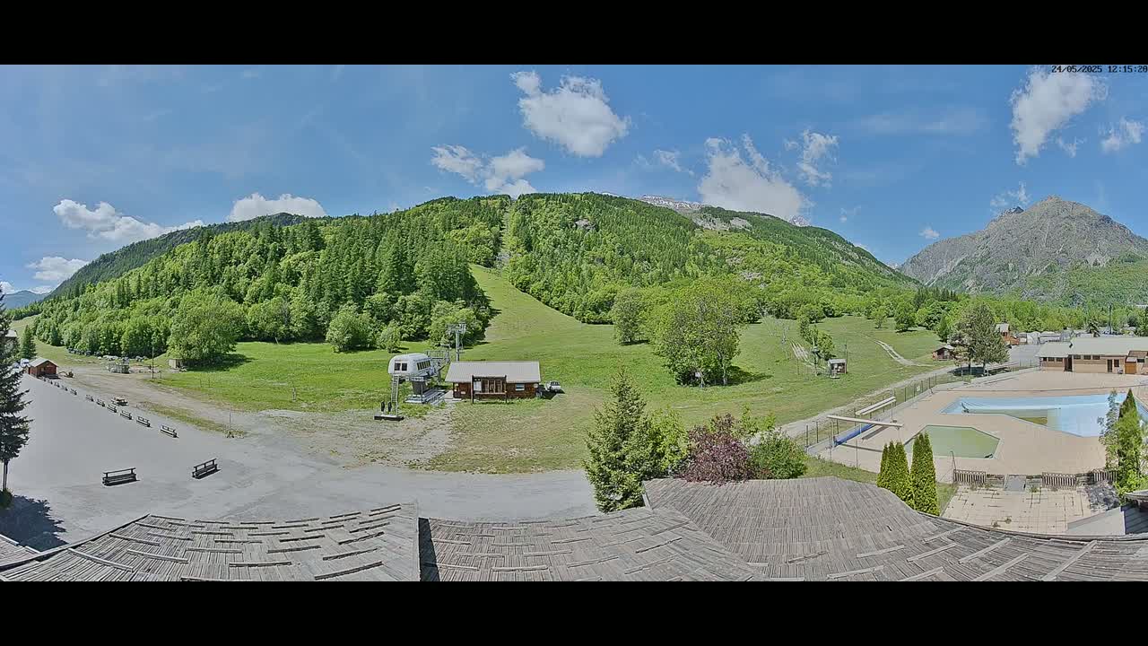 A sunny day in a mountainous region shows a grassy field with a ski lift, a small building, and an outdoor pool, all surrounded by green hills and mountains.