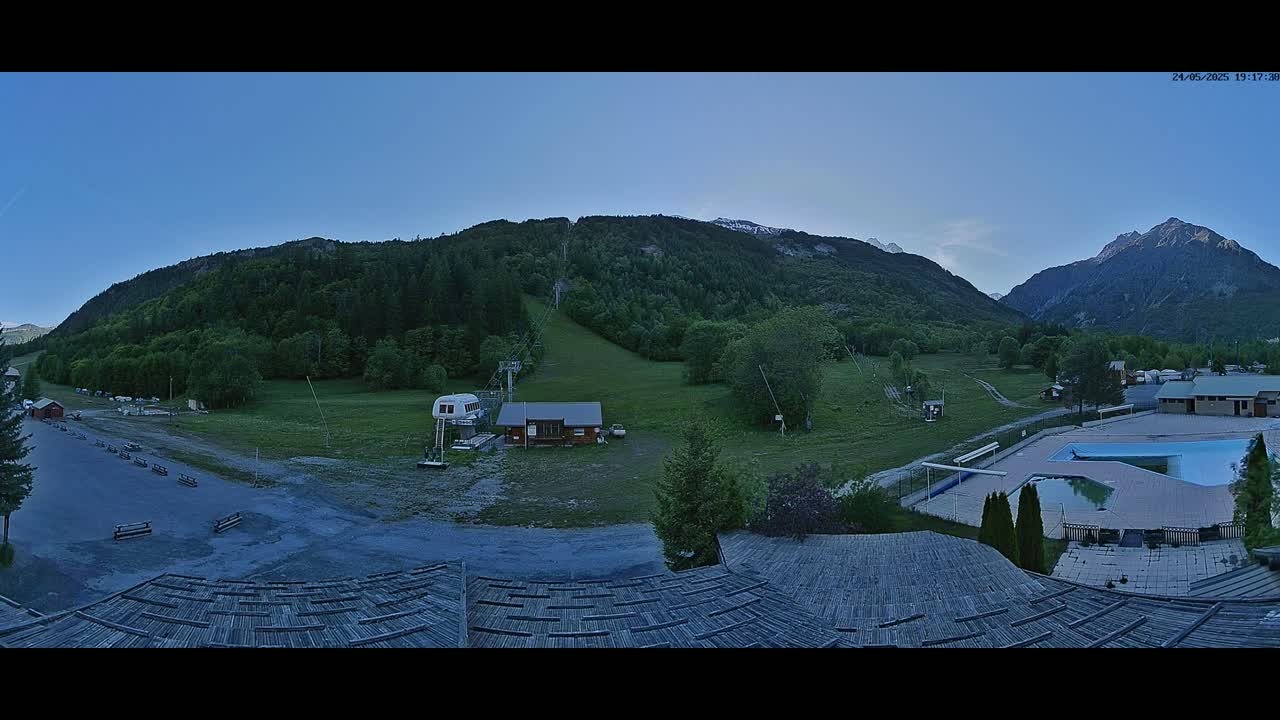 A panoramic view of a mountain valley at dusk shows a grassy field with a ski lift, a small building, a swimming pool, and forested mountains in the background under a clear sky.