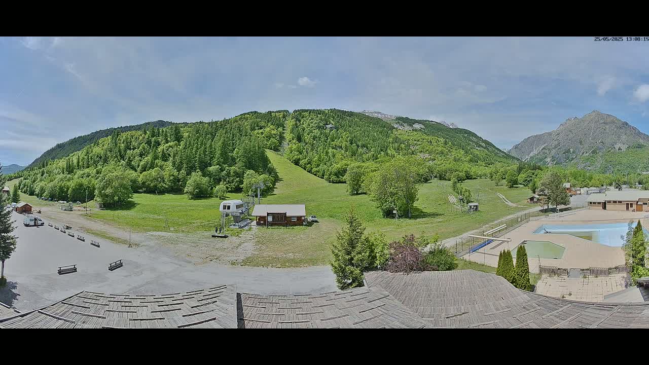 A mostly sunny day overlooking a mountain valley with green hills, a ski lift, a small building, a parking lot, and an outdoor pool.