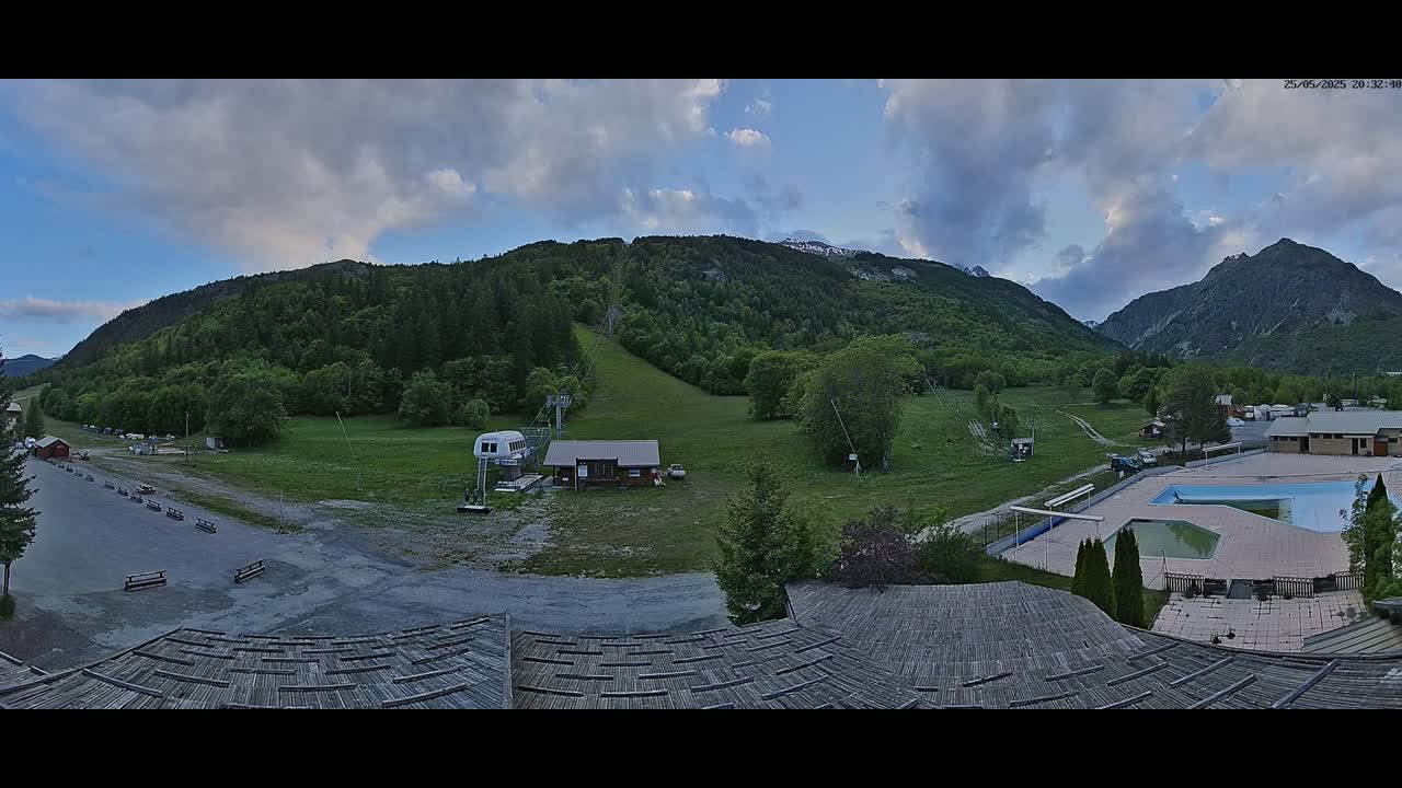 A panoramic view of a mountain valley under a partly cloudy sky shows a ski lift, a small building, a swimming pool, and a grassy area with some trees.