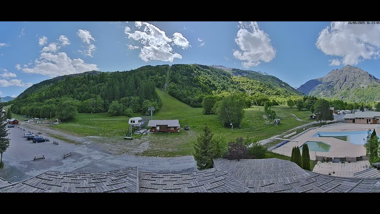 A mostly sunny day shows a mountain valley scene with a chairlift, a small building, a parking lot, and an outdoor swimming pool.