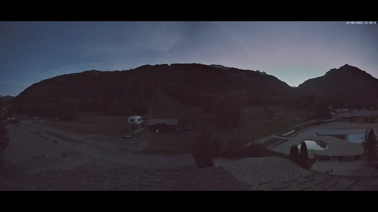 A twilight view shows a valley surrounded by dark mountains, a flat area with a few buildings and vehicles, and a swimming pool in the lower right.