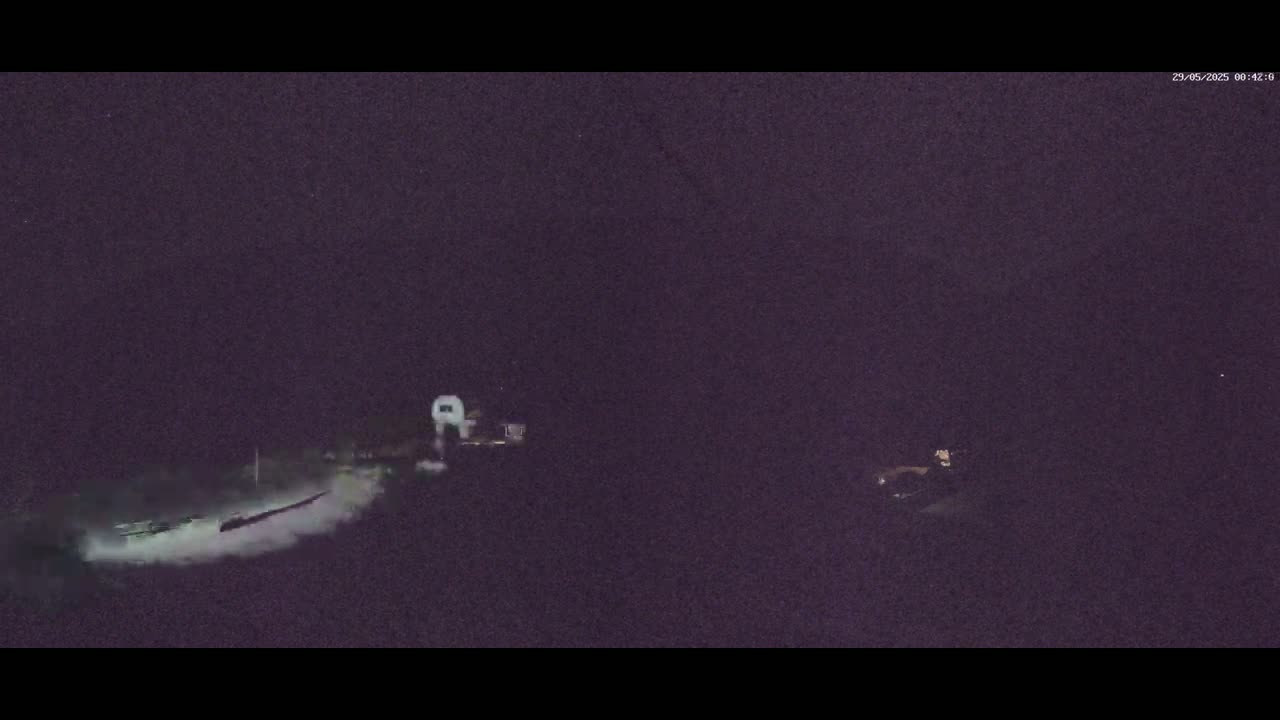 A nighttime, low-light image shows a person sitting on something near a body of water where a boat is leaving a wake.