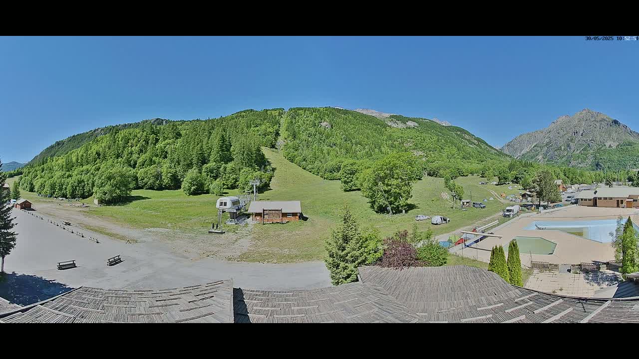 A sunny mountain valley scene with a ski lift, small buildings, a swimming pool, and green forested hills.
