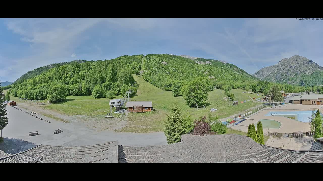 A mostly sunny view of a ski resort in the summer, showing a grassy slope, a chairlift, a small building, a parking area, and an outdoor swimming pool, all nestled in a valley surrounded by lush green mountains.