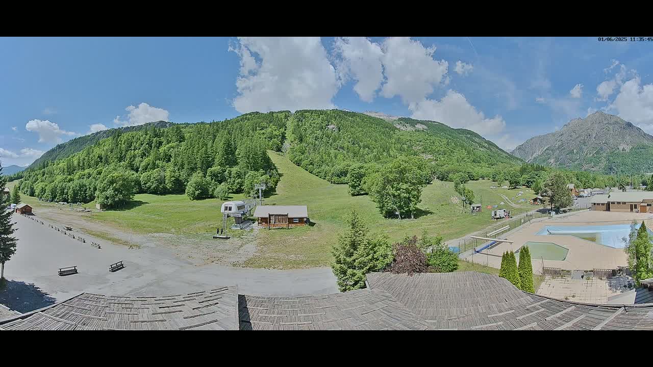 A mountain resort area with a grassy slope, a small building, a swimming pool, and surrounding green mountains under a partly cloudy sky.