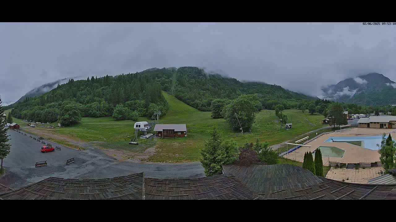 A mountainside ski resort scene on an overcast day shows a grassy slope with a chairlift, a small building, a parking lot with a few cars, and an outdoor pool, all surrounded by green hills and trees under a cloudy sky.
