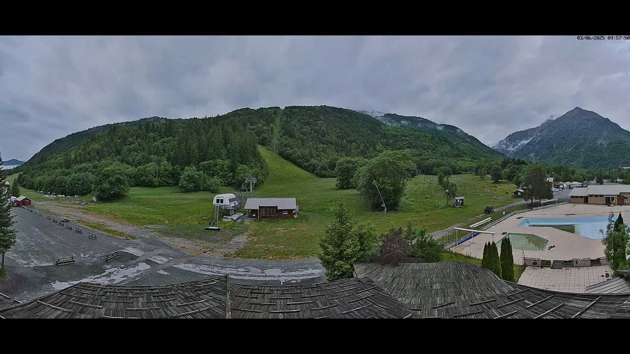 A mountain view with overcast skies shows a grassy field with a ski lift, small buildings, and an outdoor swimming pool.