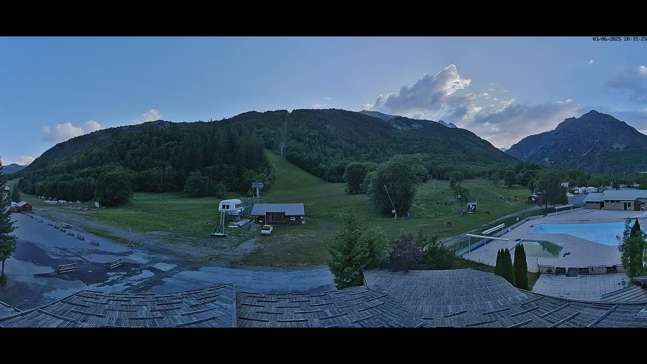 A panoramic view of a mountain valley at dusk, showing a ski lift, buildings, a swimming pool, and cattle grazing in a pasture, under a partly cloudy sky.