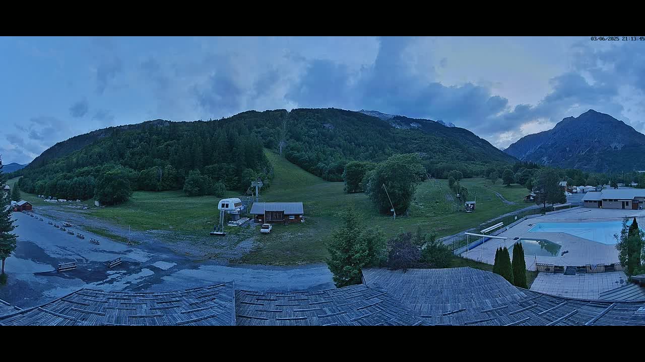 A mountain valley scene at dusk, showing a ski lift, small buildings, a swimming pool, and a large, wooded mountain range under a partly cloudy sky.
