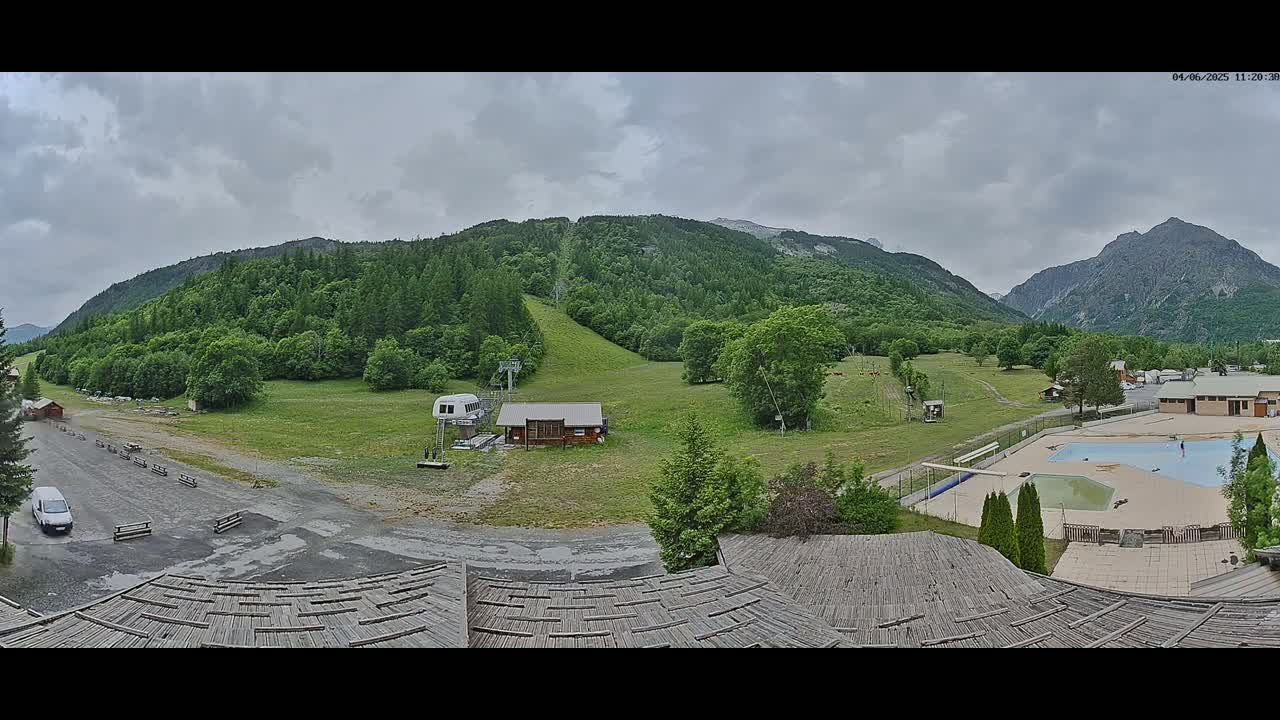 A panoramic view of a mountain valley on an overcast day shows a ski lift, grassy hillsides, a swimming pool, and several buildings.