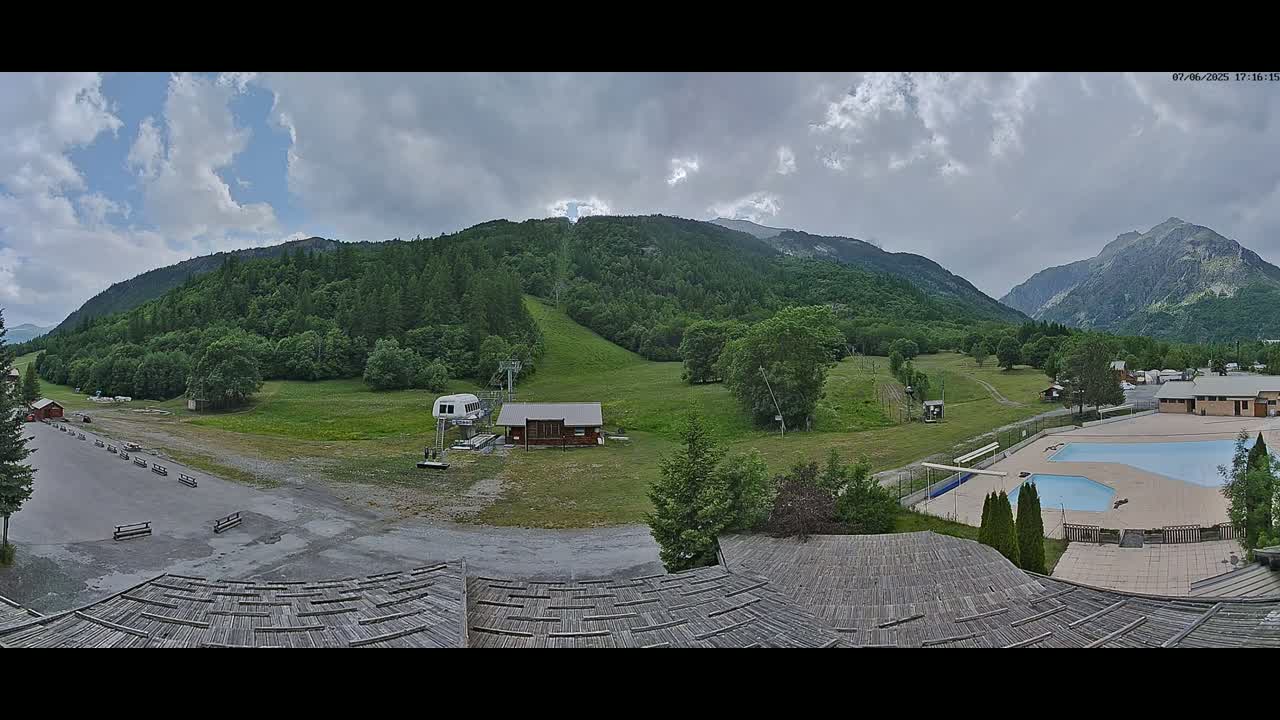 A mountain valley scene under partly cloudy skies shows a ski lift, small buildings, a swimming pool, and a large expanse of green hills and mountains.