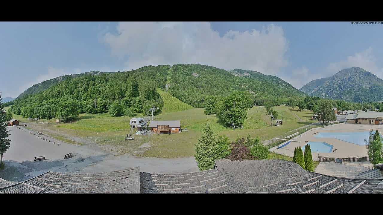 A mostly sunny day reveals a mountainous landscape with a grassy slope, ski lift, small buildings, and an outdoor swimming pool.