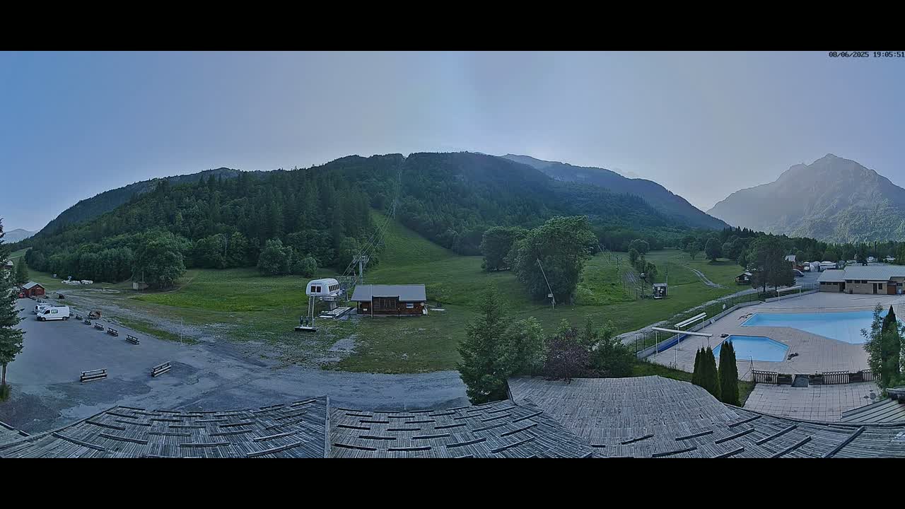A panoramic view shows a mountain valley with a ski lift, a small building, a parking lot, and an outdoor swimming pool, under a clear, light-blue sky.