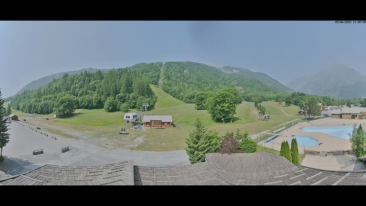 A panoramic view of a mountainside resort on a clear, sunny day shows a grassy area with a chairlift, a small building, and an outdoor swimming pool complex, all surrounded by lush green forests and mountains in the background.