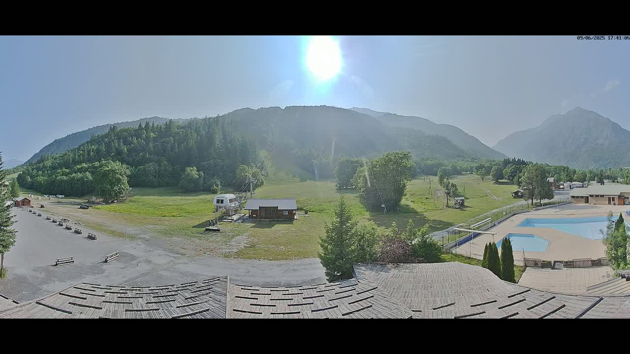 A sunny day in a mountainous region shows a grassy field with a chairlift, small buildings, a swimming pool, and a large parking lot.