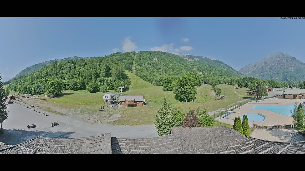 A panoramic view shows a mountainside resort area on a sunny day, featuring a grassy ski slope, a small building, a swimming pool, and a parking lot.