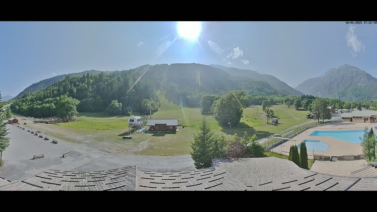 A sunny view of a mountainous landscape with a grassy field, a small building, a swimming pool, and a ski lift in the distance.