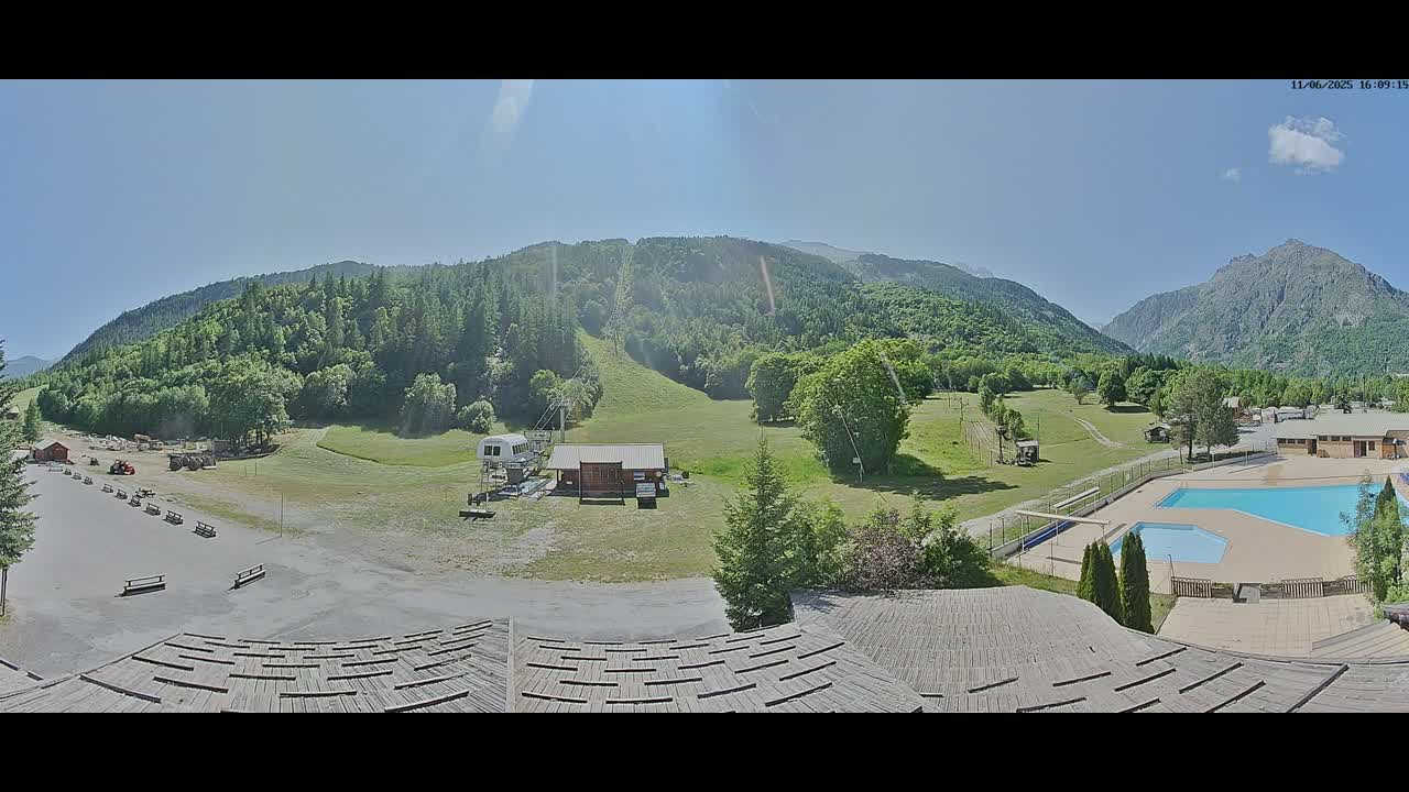 A sunny outdoor view shows a mountain valley with a grassy area, a ski lift, a building, a swimming pool, and a parking lot.