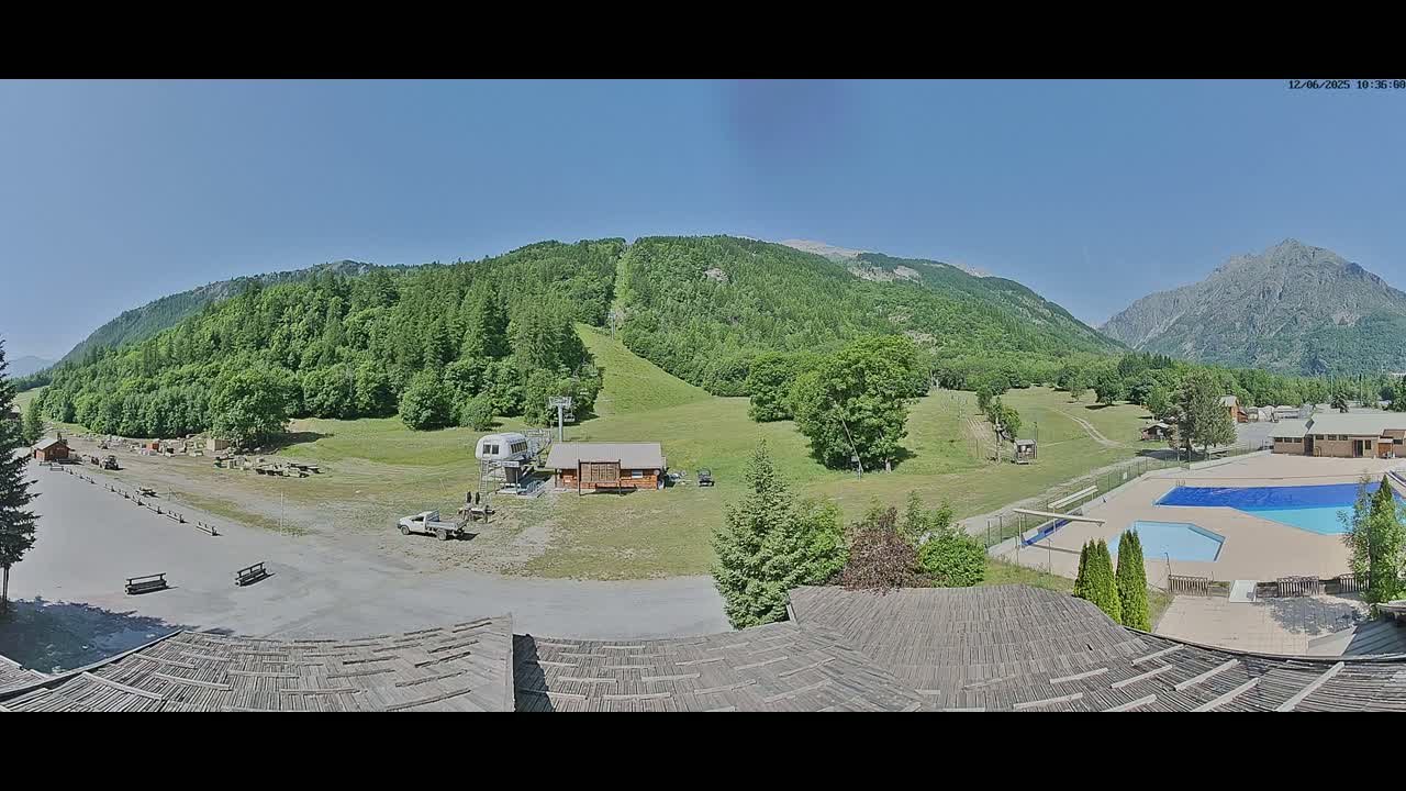 A mountain resort on a sunny day with a parking lot, a small building, a chairlift, a swimming pool, and lush green mountains in the background.